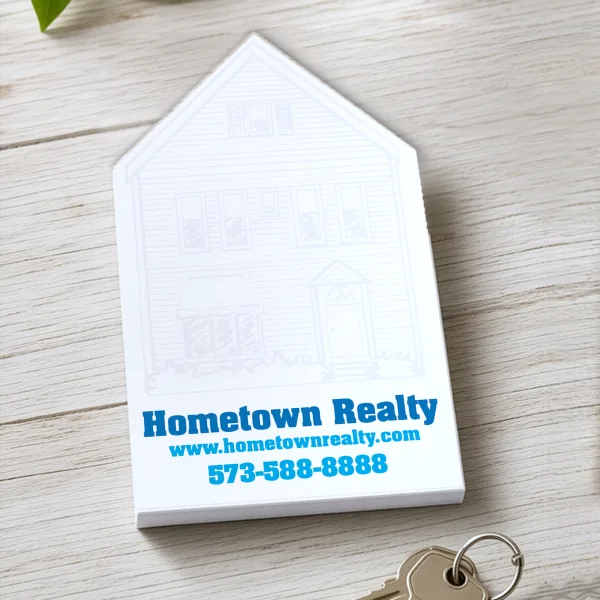 House-shaped die cut Hometown Realty notepad with a sketched home, on light wood beside house keys and a green leaf.