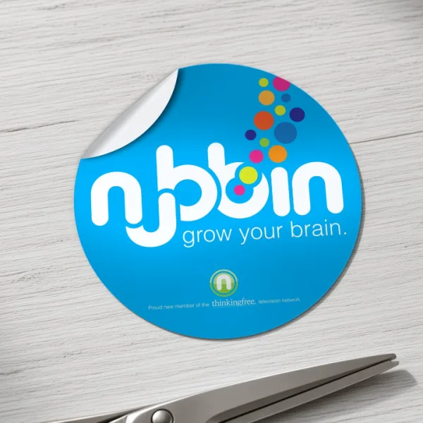 Round custom nubbin "grow your brain" sticker with a peeled-back corner on light wood, beside a pair of scissors.