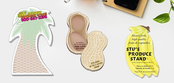 Three die-cut shaped magnets including a green palm tree, a tan peanut, and a yellow banana on a white desk surface.