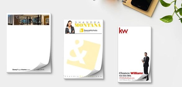 Three real estate notepads with agent photos and branding featuring sticky tabs on a white desktop with phone.
