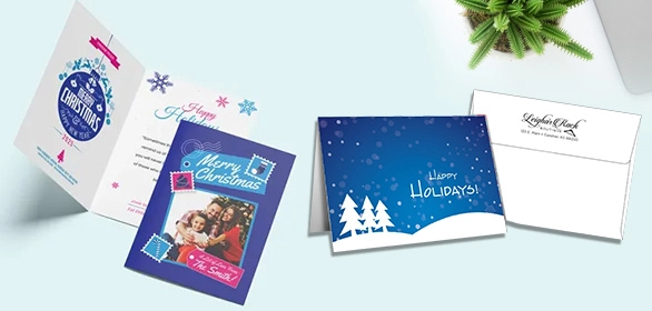 Folded holiday greeting cards with snowflakes, Christmas trees, family photo, and a white envelope near a green fern.