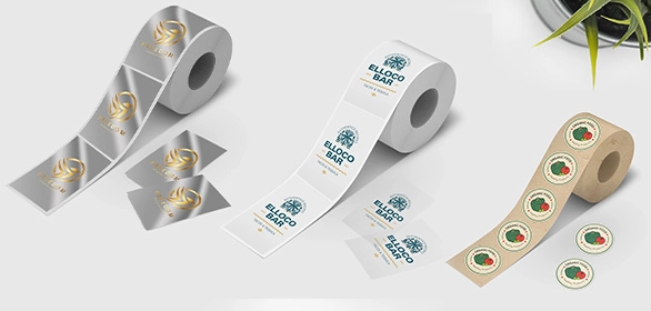Three rolls of adhesive labels in silver, white, and kraft brown with printed logos on a white surface near a plant.