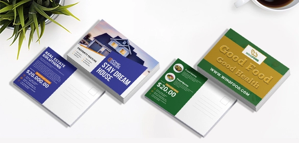 Two stacks of postcards featuring a blue house design and a green and gold food-themed layout on a desk.