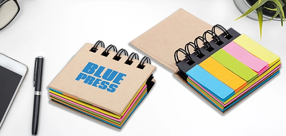 Two kraft spiral bound mini notebooks with colored sticky note tabs beside a pen and phone on white.