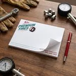 Custom 6 x 4 inch Premium Tires notepad flat on a workbench with gloves, lug nuts, tire gauges and a red pen