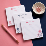 Custom 4 x 4 inch Aster Lane square pads, three checkbox pads with flowers, a pink pen and a dish of paper clips